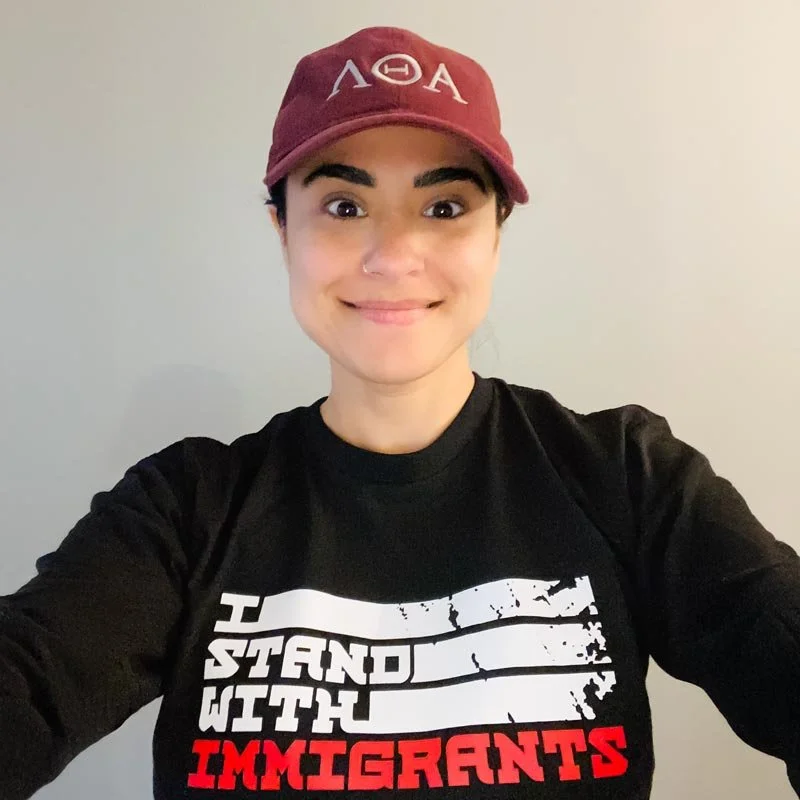 A young woman wearing a maroon baseball cap with Greek letters and a black long-sleeve shirt that reads "I stand with immigrants" in white and red text, smiling at the camera against a plain light-colored background.
