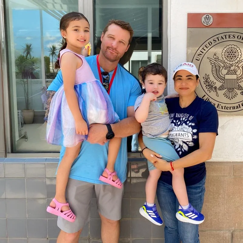 A family of four, including two young children, stands in front of a government building with a U.S. Department of Homeland Security sign. The father holds a girl in a colorful dress, and the mother holds a boy in a gray shirt. All are smiling.