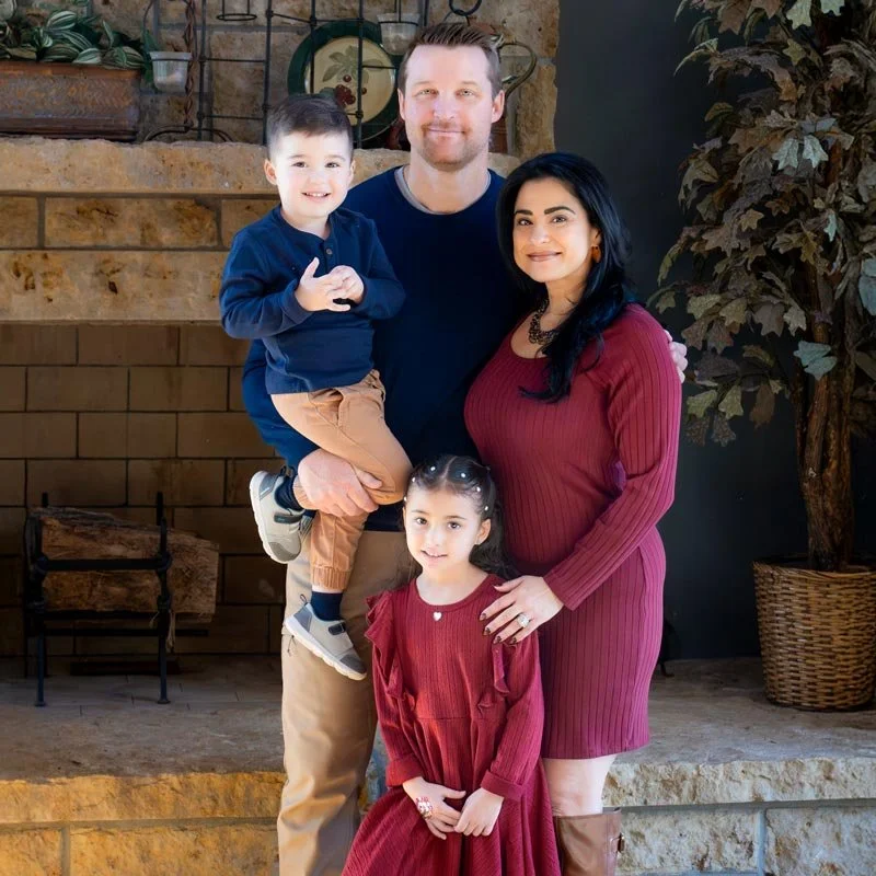A family of four posing indoors near a stone fireplace. The father holds a young boy, the mother stands beside a young girl, all smiling.