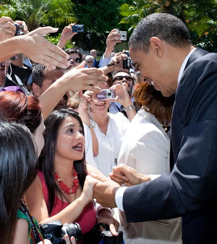Man in a dark suit smiling and holding hands with a woman in a red top, in front of a crowd taking pictures with cameras and smartphones.