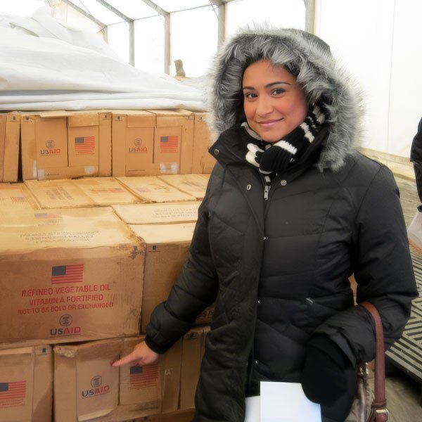 Woman in a black winter coat with a fur-lined hood and scarf standing next to boxes of U.S. Aid supplies in a warehouse.