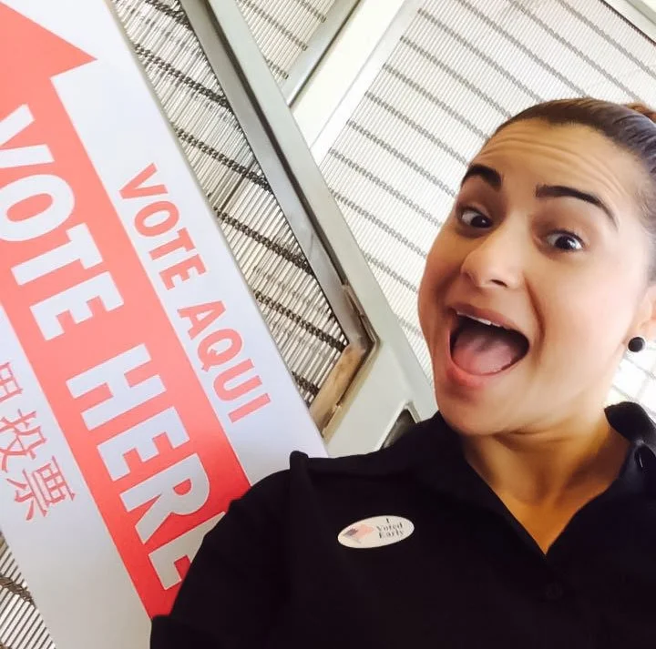 A woman smiling in a black shirt with an "I voted pin." Behind her is a "Vote Here" sign. 