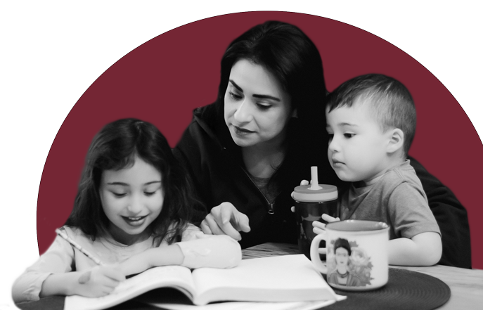 A woman with two young children reading a book together at a table, with a mug and a sippy cup on the table, against a maroon background.