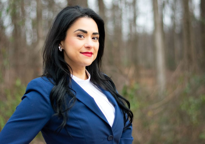 A woman with black hair, wearing a blue blazer and white blouse, standing outdoors in a wooded area with trees in the background.