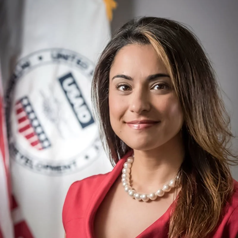 A woman with long brown hair and a pearl necklace, wearing a red blazer, smiling at the camera, with a blurred background featuring an American flag and a logo.
