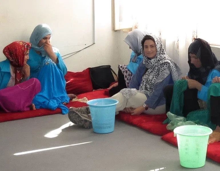 Group of women and girls sitting on a red carpet in a room, some wearing headscarves, engaged in a craft activity.