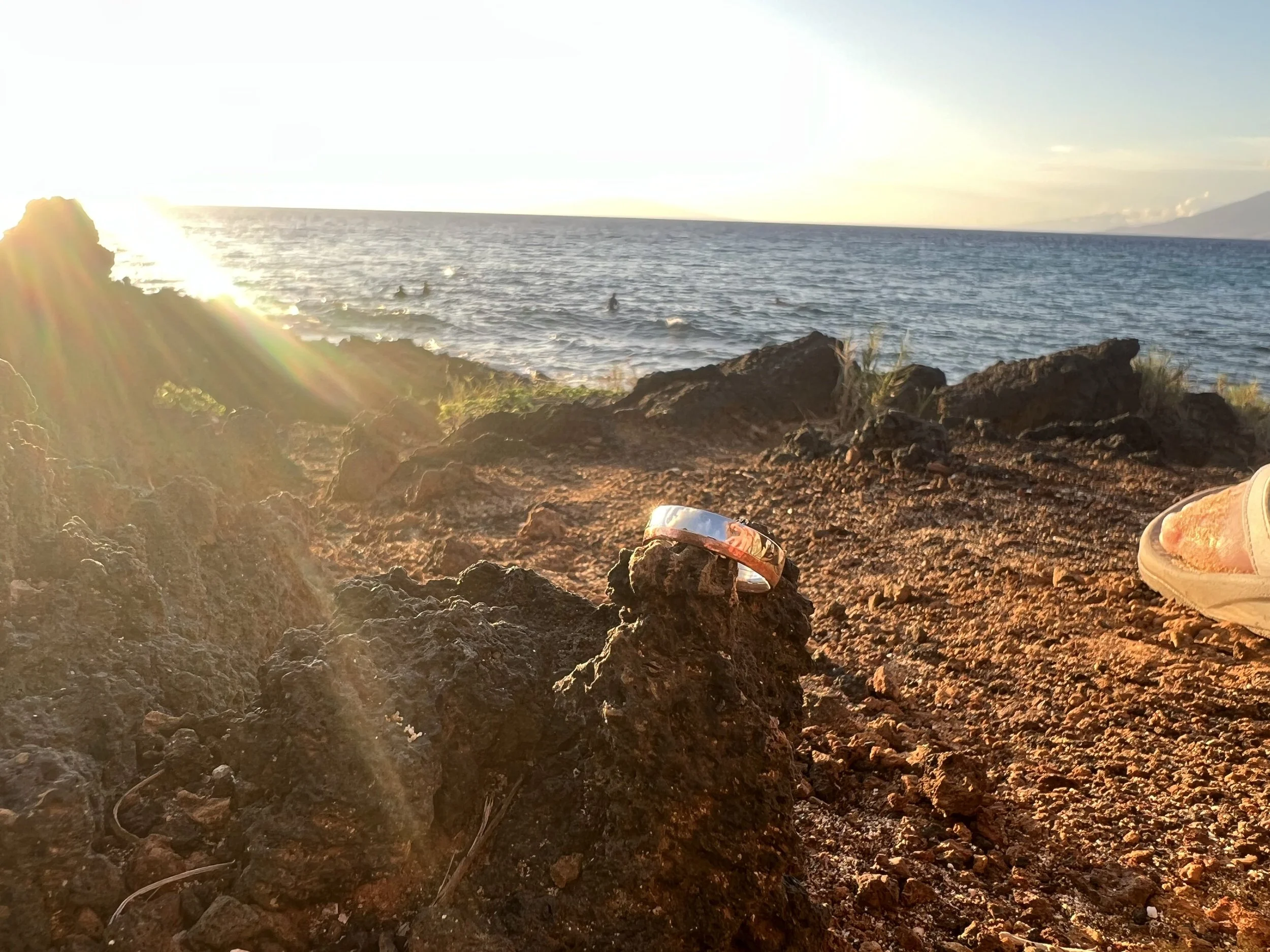 A rocky coastline with the ocean in the background, sunlight creating lens flare, and a silver bracelet resting on a rock in the foreground. Part of a person's foot is visible on the right side.