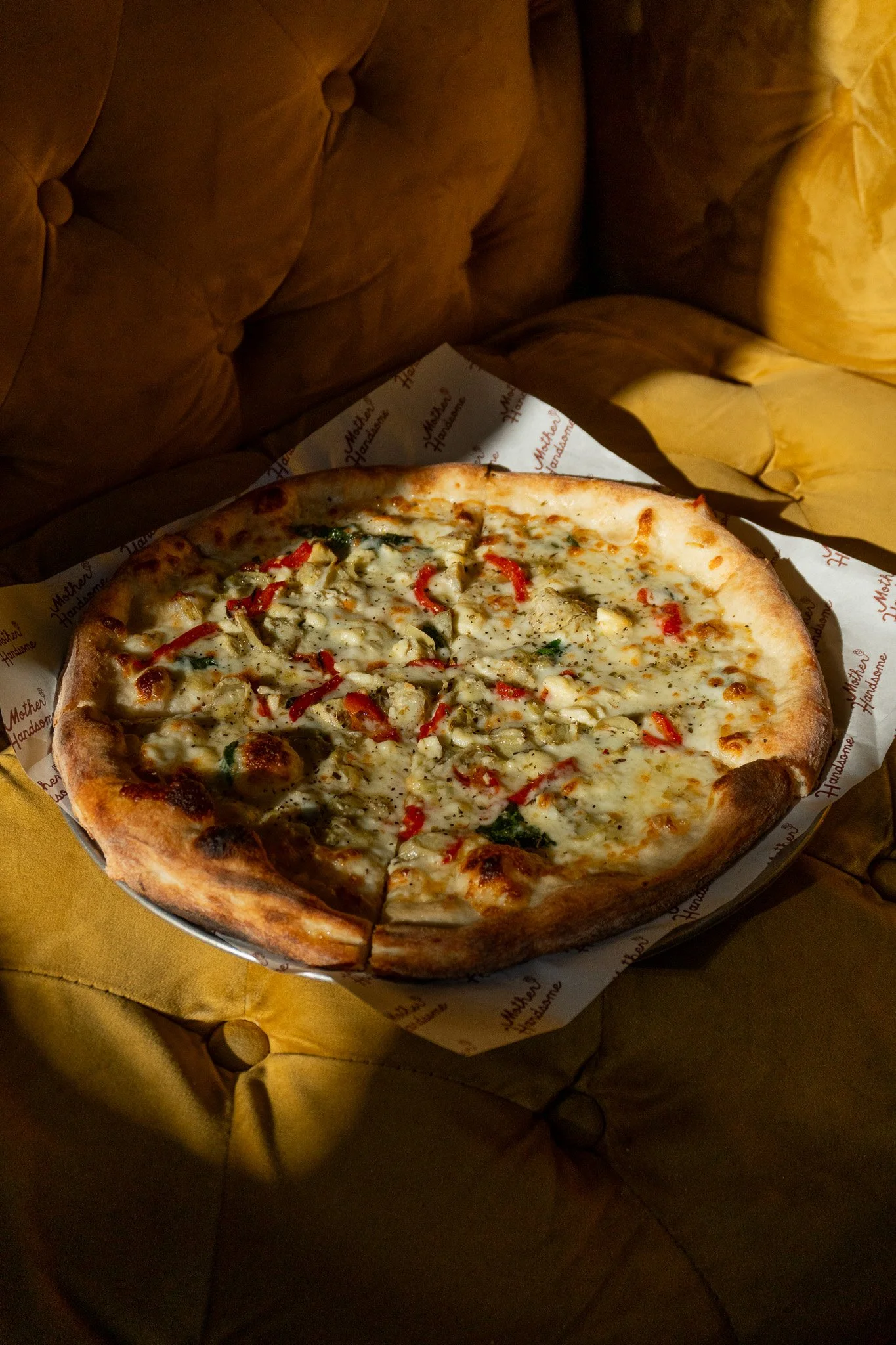 A pizza with cheese, red peppers, green herbs, and possibly chicken or chicken sausage, on a paper-lined tray.