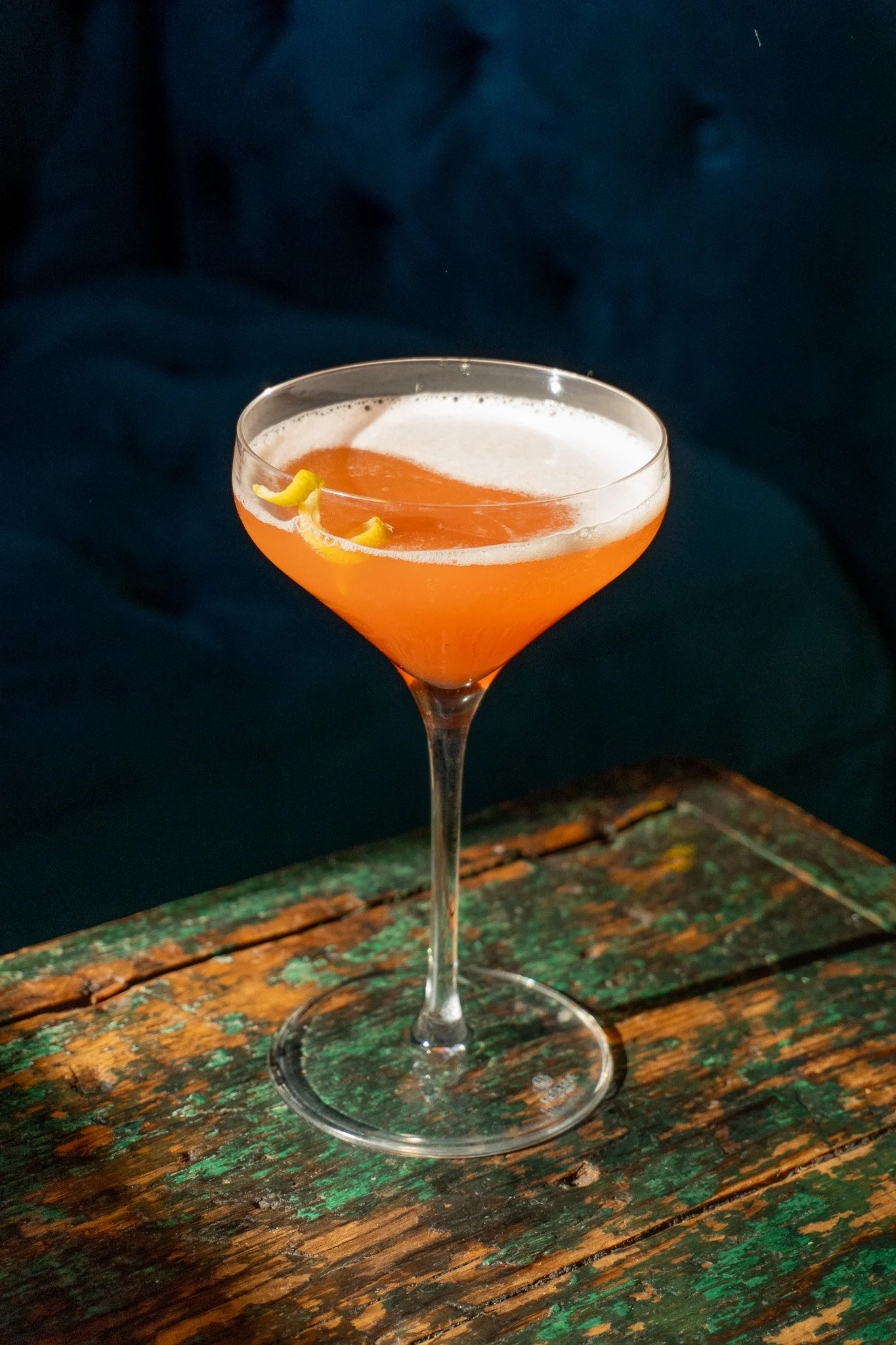 A cocktail in a coupe glass with an orange-colored drink and a lemon peel garnish, set on a rustic wooden surface with a dark background.