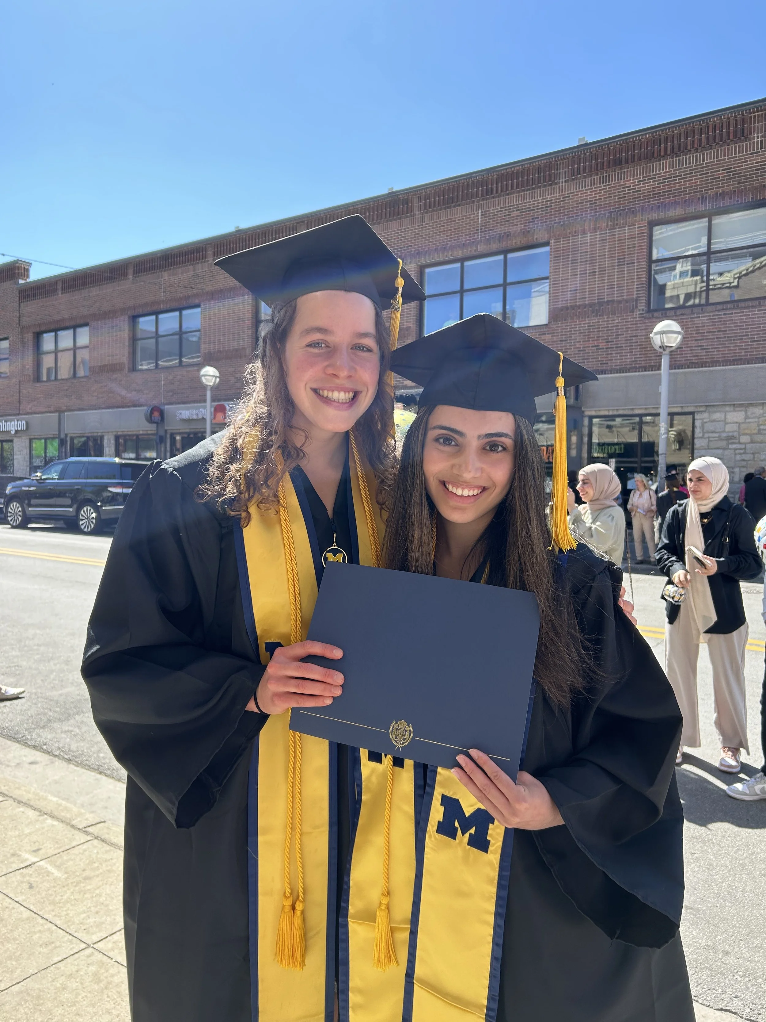Two young women in graduation caps and gowns celebrating graduation outside on a sunny day, one holding a diploma.