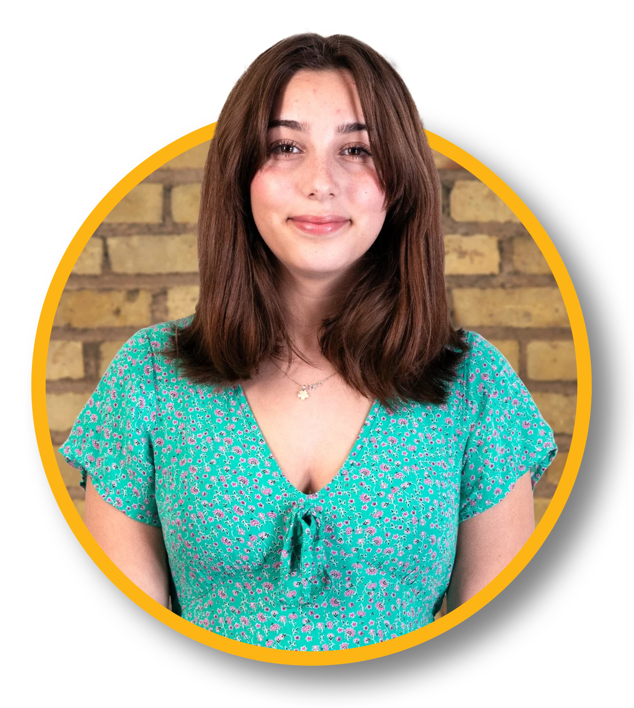A woman with shoulder-length brown hair, wearing a teal floral dress and a necklace, smiling in front of a brick wall.