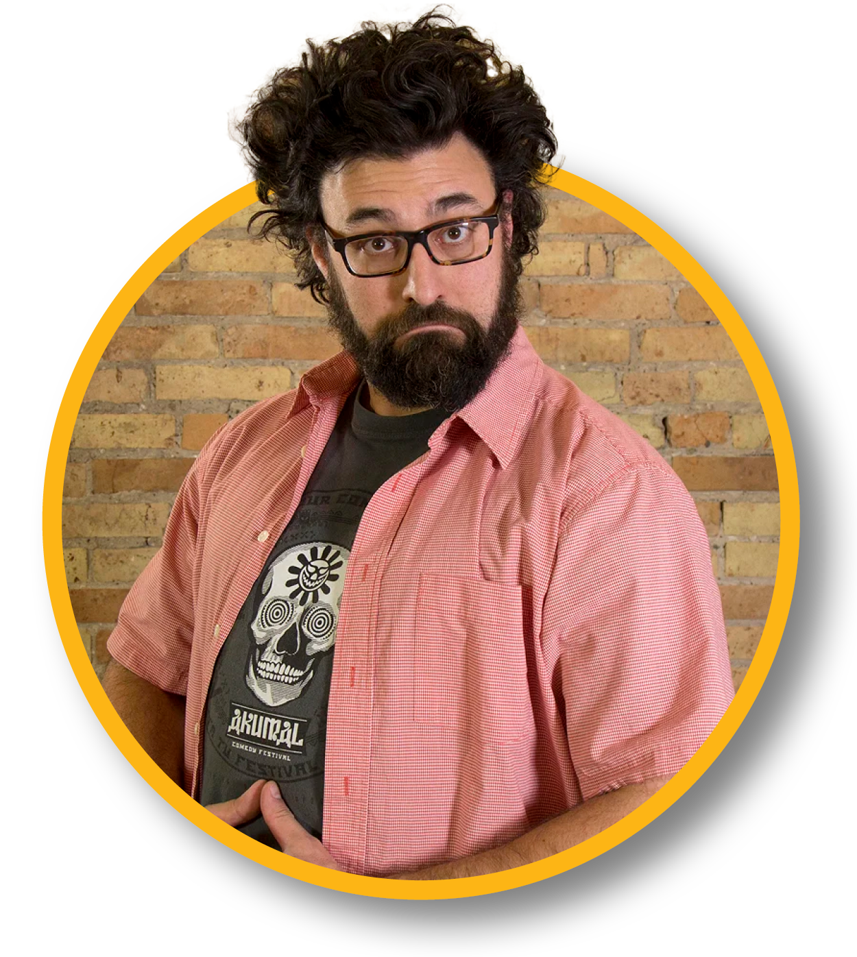 A man with curly dark hair, glasses, and a beard, standing against a brick wall. He is wearing a pink short-sleeved button-up shirt over a black shirt with a skull graphic and stylized text.