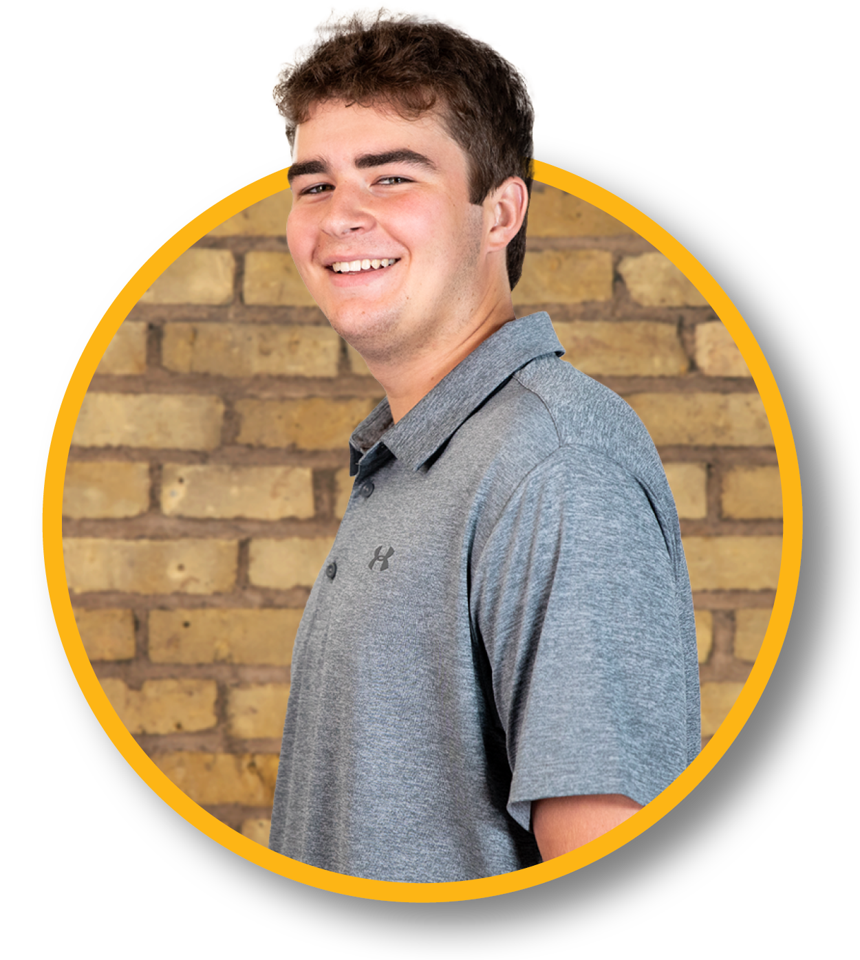 A young man smiling, wearing a gray polo shirt with an Under Armour logo, standing against a brick wall.