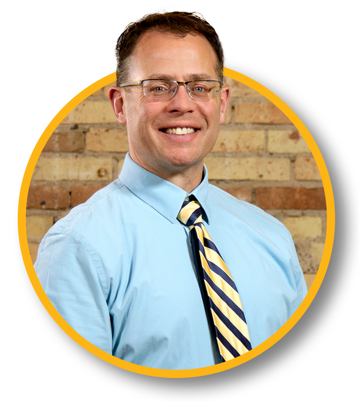 A smiling man wearing glasses, a light blue shirt, and a navy and yellow striped tie, standing in front of a brick wall.
