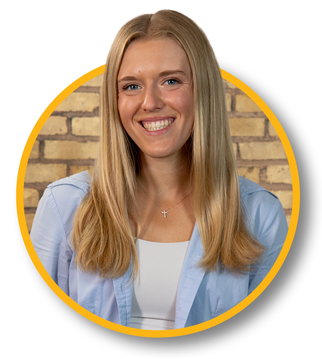 A smiling woman with long blonde hair, wearing a light blue shirt and a cross necklace, is sitting in front of a brick wall.