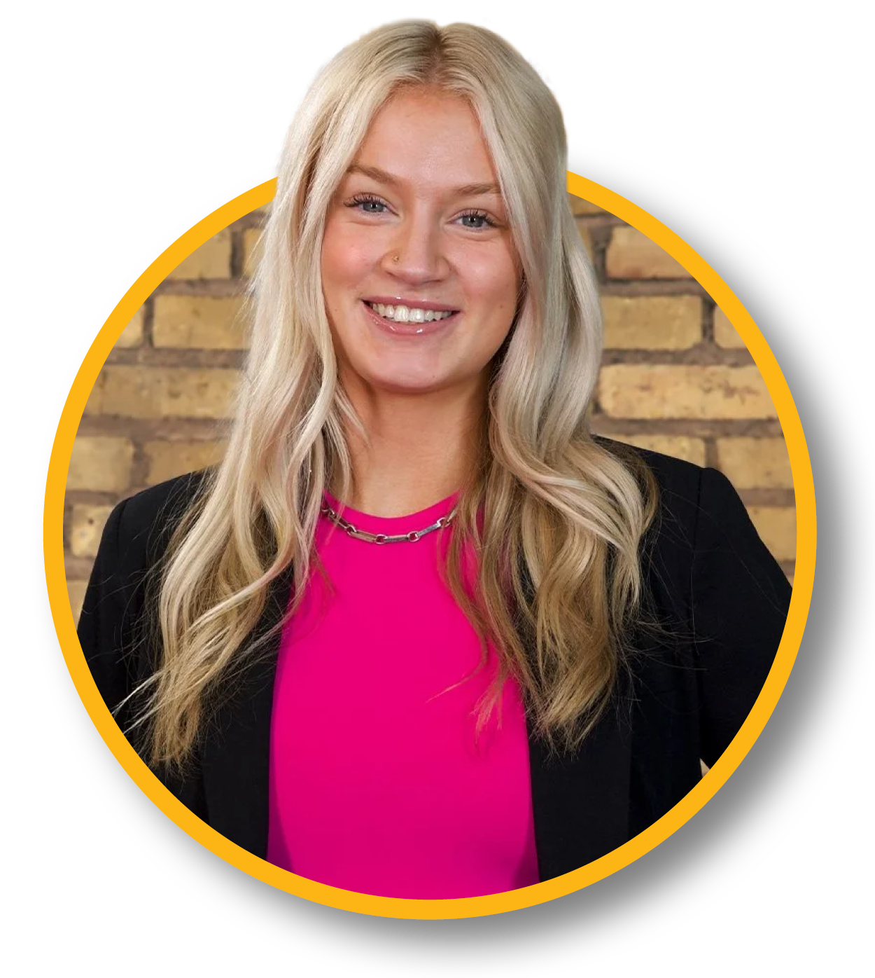 A smiling young woman with long blonde hair, wearing a black blazer and a bright pink top, standing against a brick wall background.