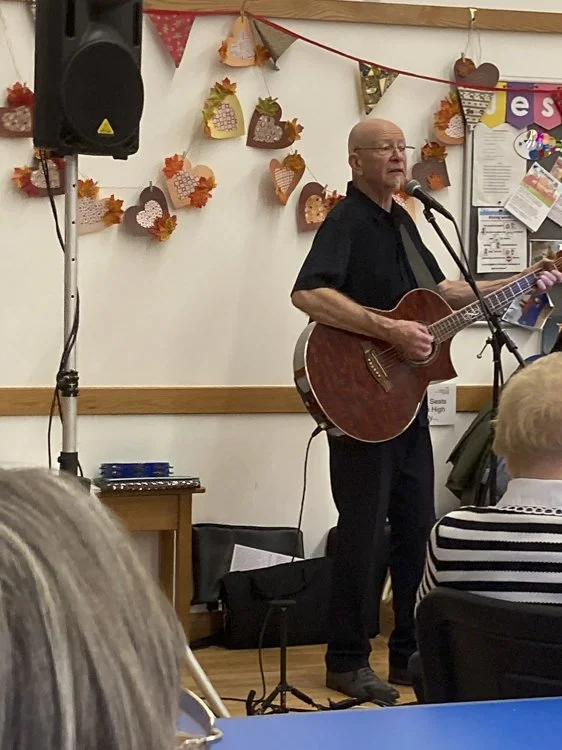 A man playing an acoustic guitar and singing into a microphone during a performance at a community event. Decorations of autumn leaves and hearts are on the wall behind him.