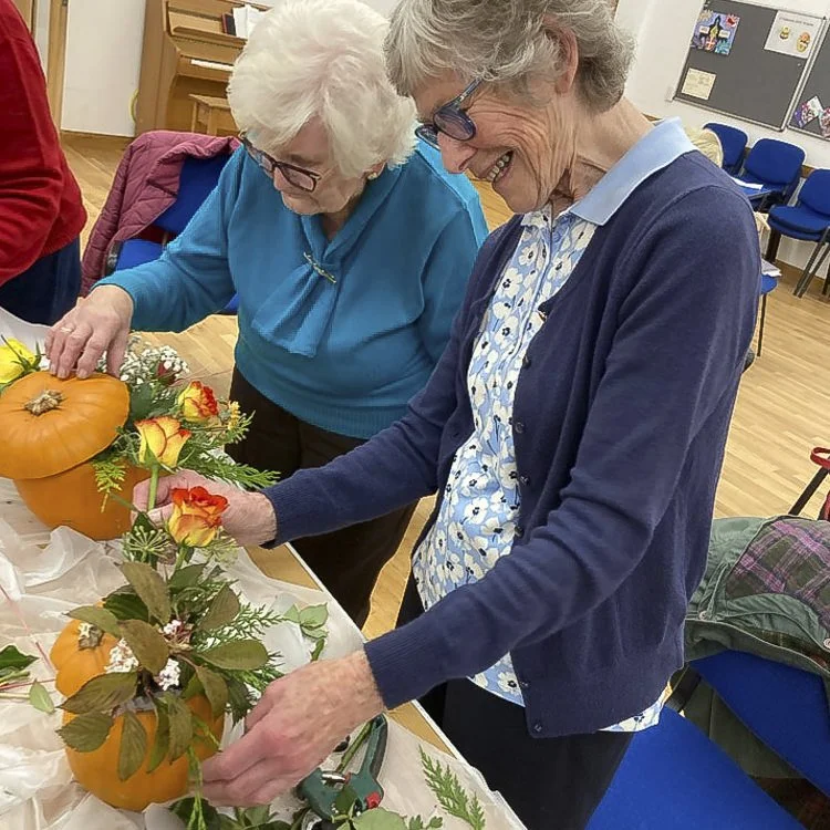 Two elderly women decorating pumpkins with flowers and foliage at a table in a community hall.