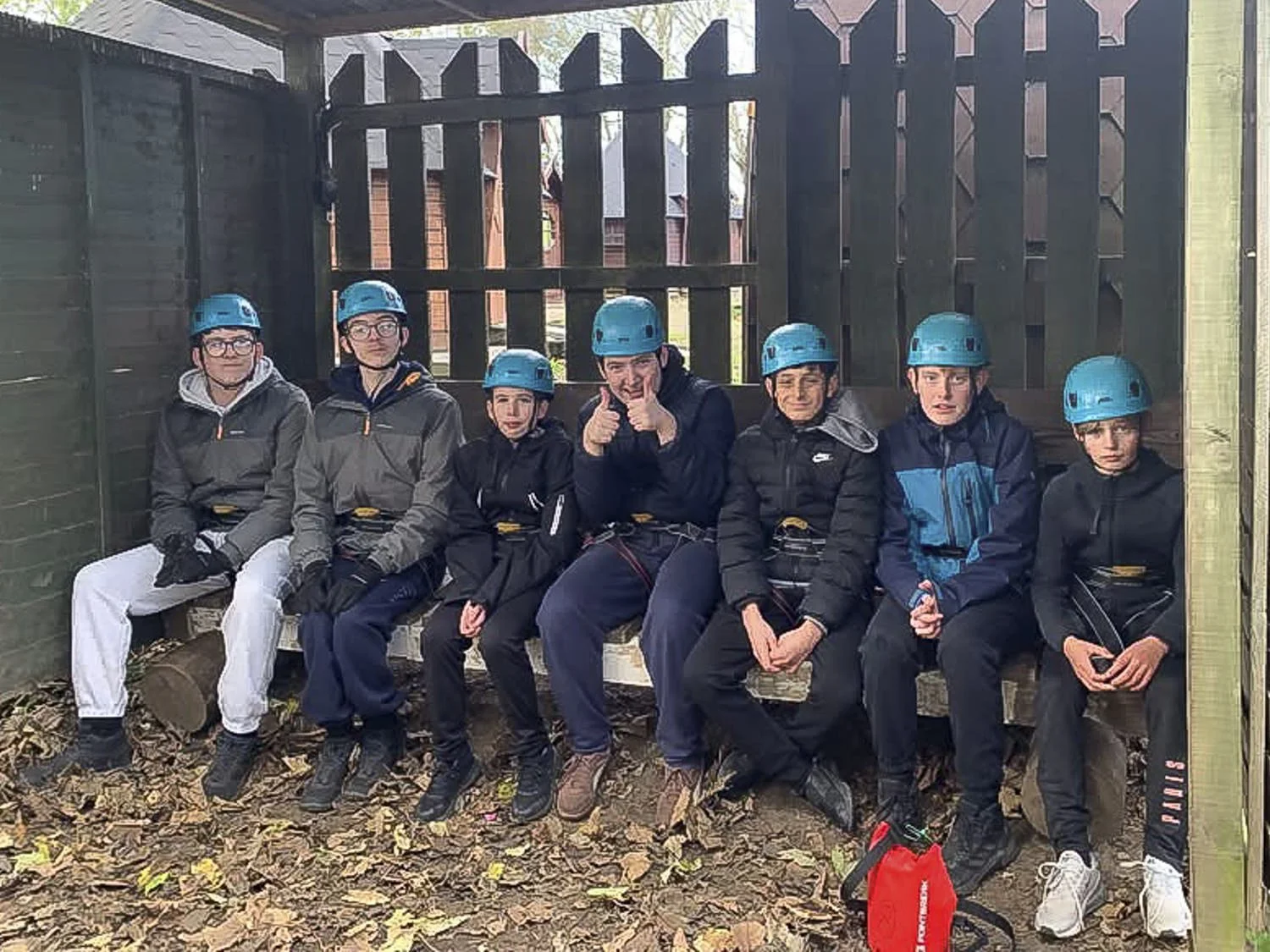 Group of seven boys wearing helmets sitting on a wooden bench inside a wooden shelter, surrounded by fallen leaves on the ground.