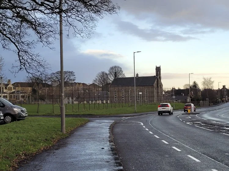A curved road with cars, a pavement, a church, and houses in the background, with a partly cloudy sky.