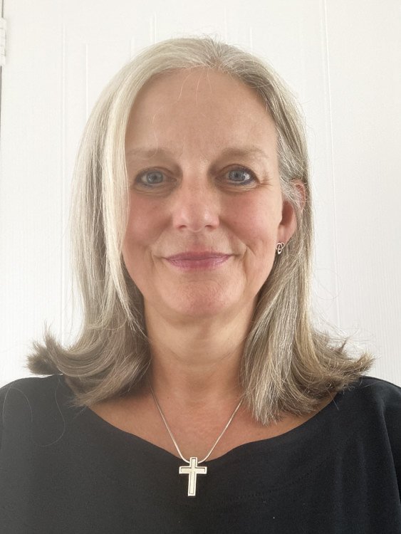 A woman with shoulder-length grey hair and blue eyes, smiling, wearing a black top, a silver cross necklace, and small earrings..