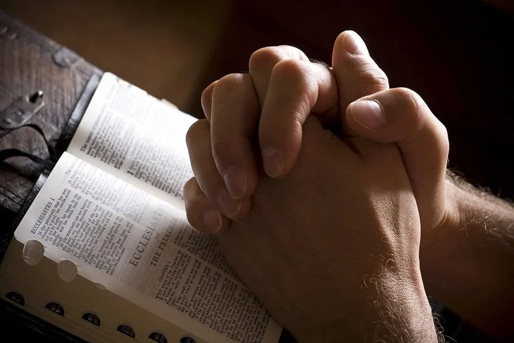 A person praying with hands clasped in front of an open Bible.