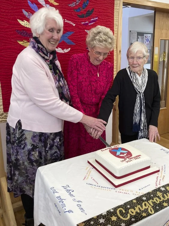 Three elderly women are gathered around a table with a cake, cutting it together. They are smiling and appear happy, in front of a red quilt with Guild logo.