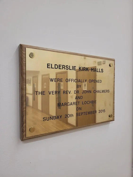 Plaque on the wall with engraved text about the opening of Elderslie Kirk Halls by Reverend Dr. John Chalmers and Margaret Lochrie on September 20, 2015, with a background of a hallway in a building.