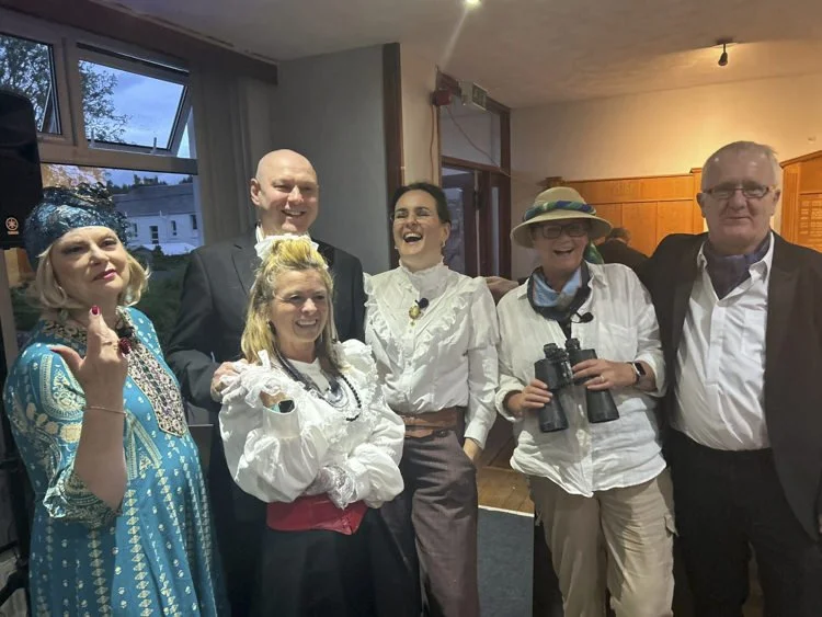 Six people dressed in vintage or costume clothing, standing indoors in a room with wooden paneling and a window. One woman is wearing a blue dress and headscarf, making a rock gesture. Another woman is in a white blouse and black skirt, smiling. The man next to her is in a suit, smiling. The woman in the middle is dressed in a white blouse and brown skirt, laughing. The woman beside her is wearing a beige hat, glasses, and holding binoculars. The man on the far right is in a white shirt and dark jacket, with his arm around the woman with binoculars.