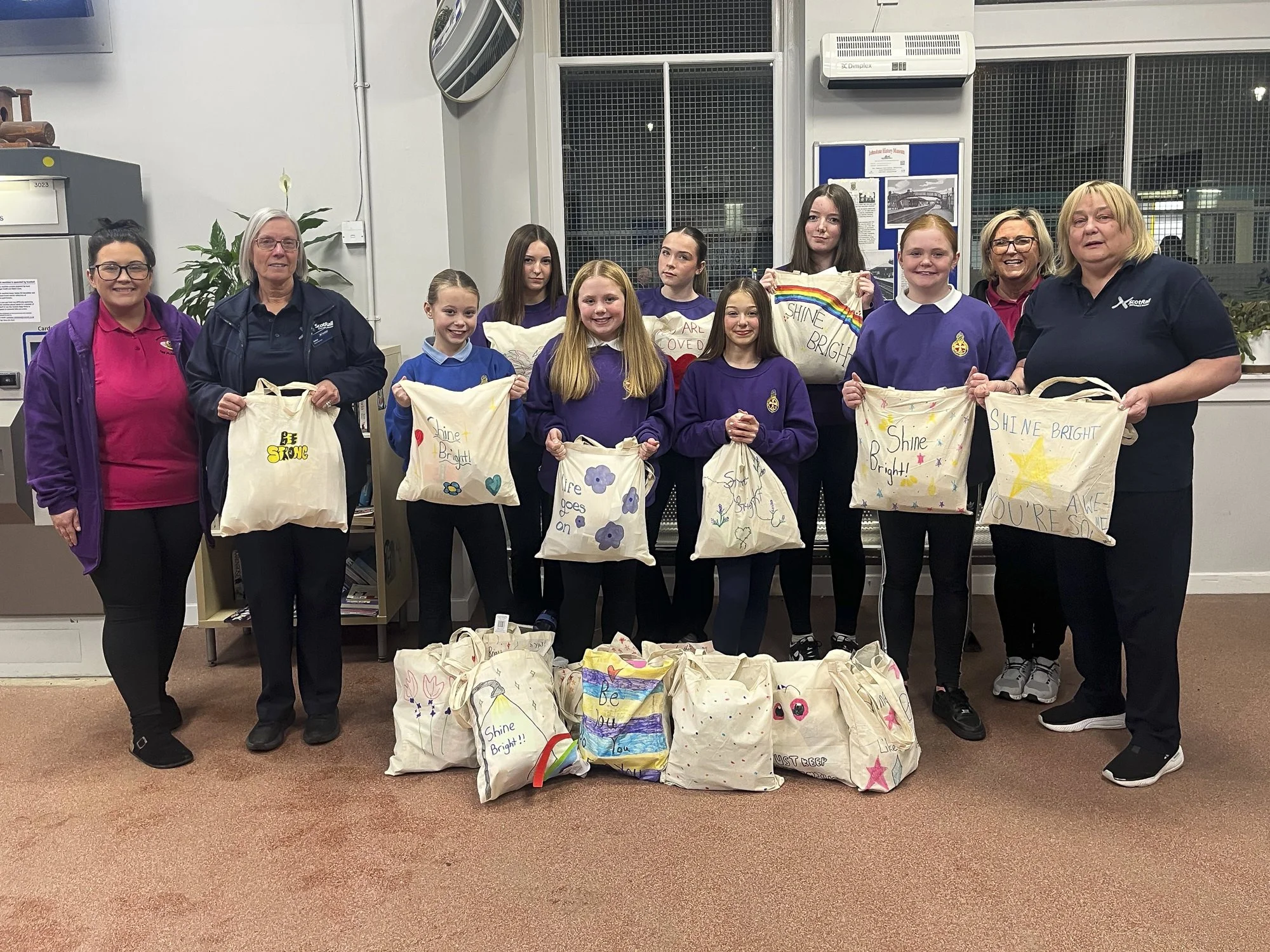 Group of people, including girls and adults, standing indoors holding handmade tote bags with colourful messages and drawings for charity.