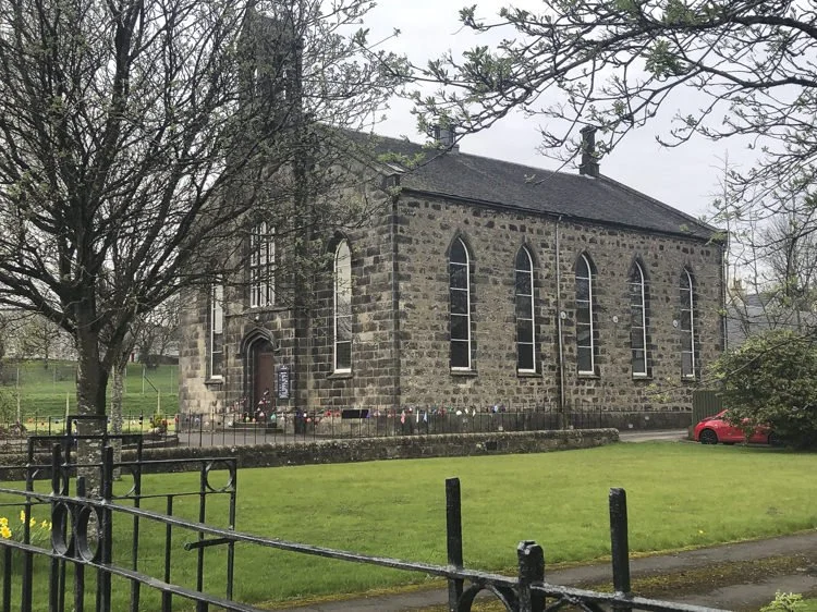 A stone church with tall arched windows, surrounded by a grassy area, trees, and a black wrought iron fence.