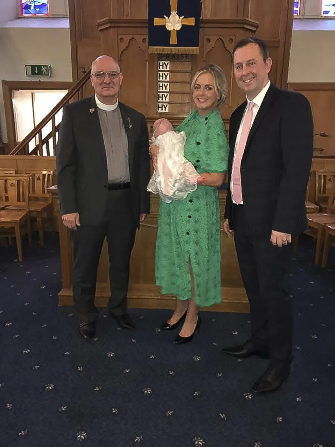 A couple with a baby in baptism outfit stand beside a minister in front of the pulpit of a church.