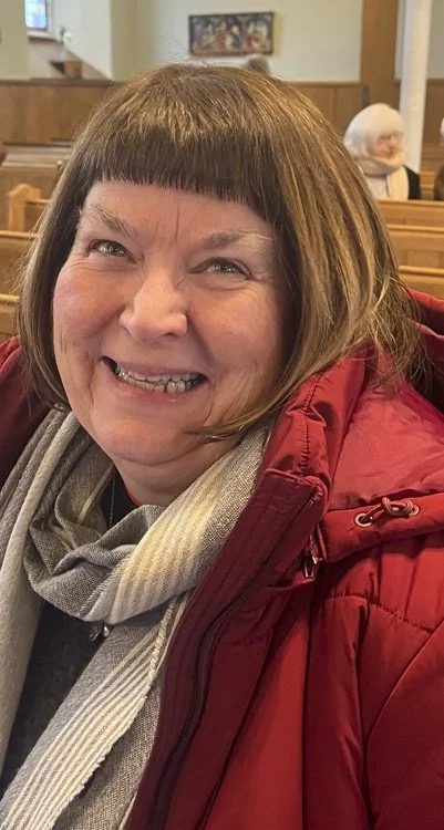 A cheerful woman with short brown hair, wearing a red jacket and a scarf, smiling inside a church with wooden pews.