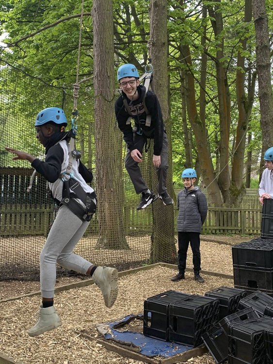 Teenagers wearing helmets enjoying an outdoor ropes course among trees.