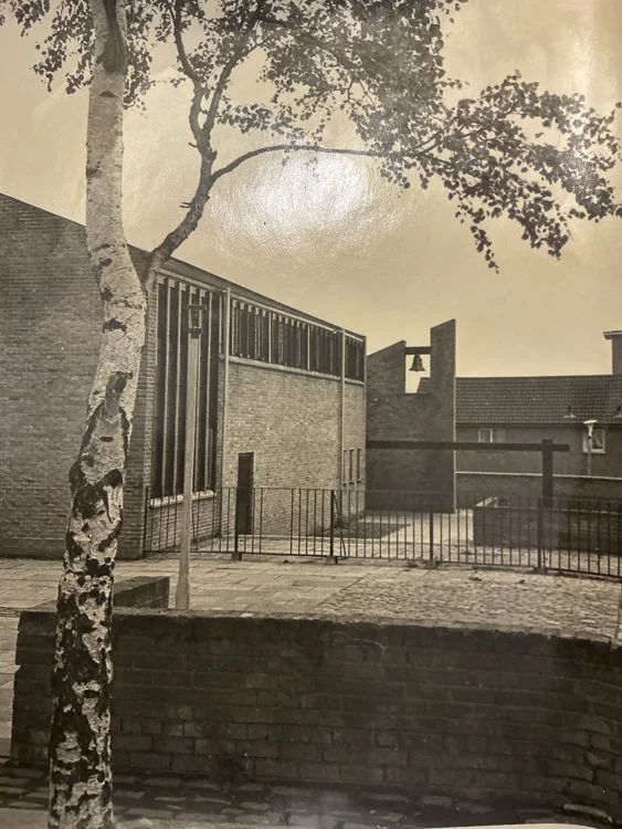 Black and white photo of a courtyard with a tree in the foreground, a building with large windows, brick walls, and a gated area.