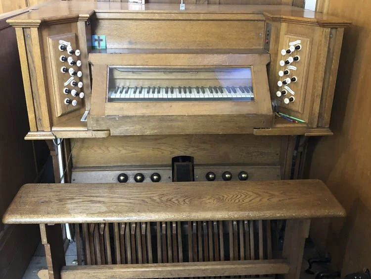 A church wooden pipe organ with a glass-covered keyboard, various stops and knobs, and a wooden bench in front.