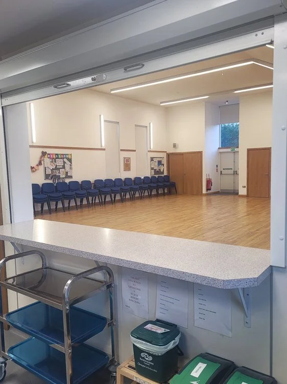 Large church hall with blue chairs along the wall, wood flooring, and a small bulletin board. Viewing from a kitchen service hatch with a countertop, shelving, and recycling bins in the foreground.