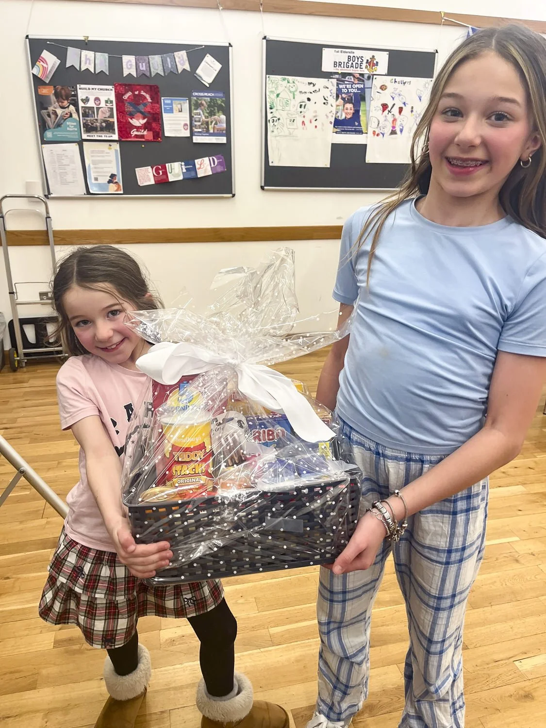Two young girls are smiling. Together they carry a big gift wrapped box containing sweets and treats.