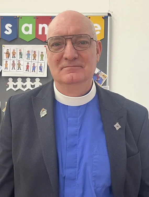 A man in a grey suit and clerical collar, wearing glasses, standing in front of a bulletin board with colourful signs and drawings.