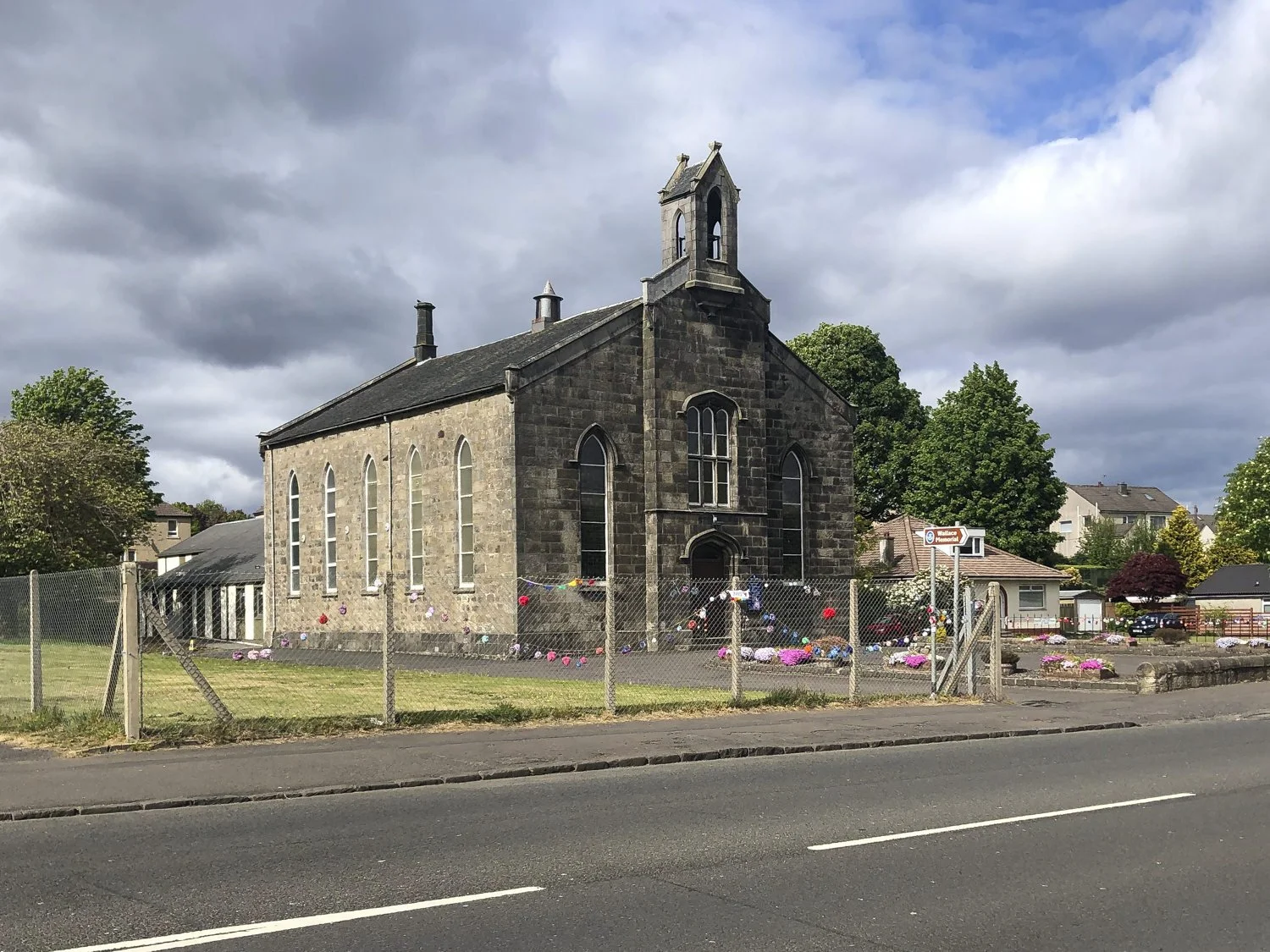 A stone church with a small bell tower and arched windows, surrounded by a chain-link fence decorated with pink, purple, and red flowers, with cloudy skies overhead.