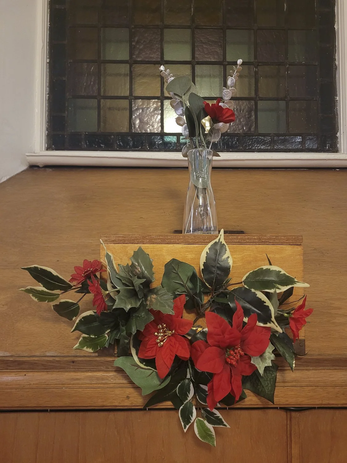 A red and green poinsettia floral arrangement and a glass vase containing red flowers and greenery in front of a stained glass window.