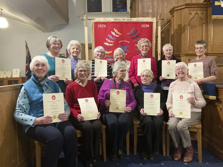 A group of eleven senior women holding certificates, posing in a church with a red memorial banner behind them that reads '1924 - 2024' and features a dove and the words 'whose, we are...'.