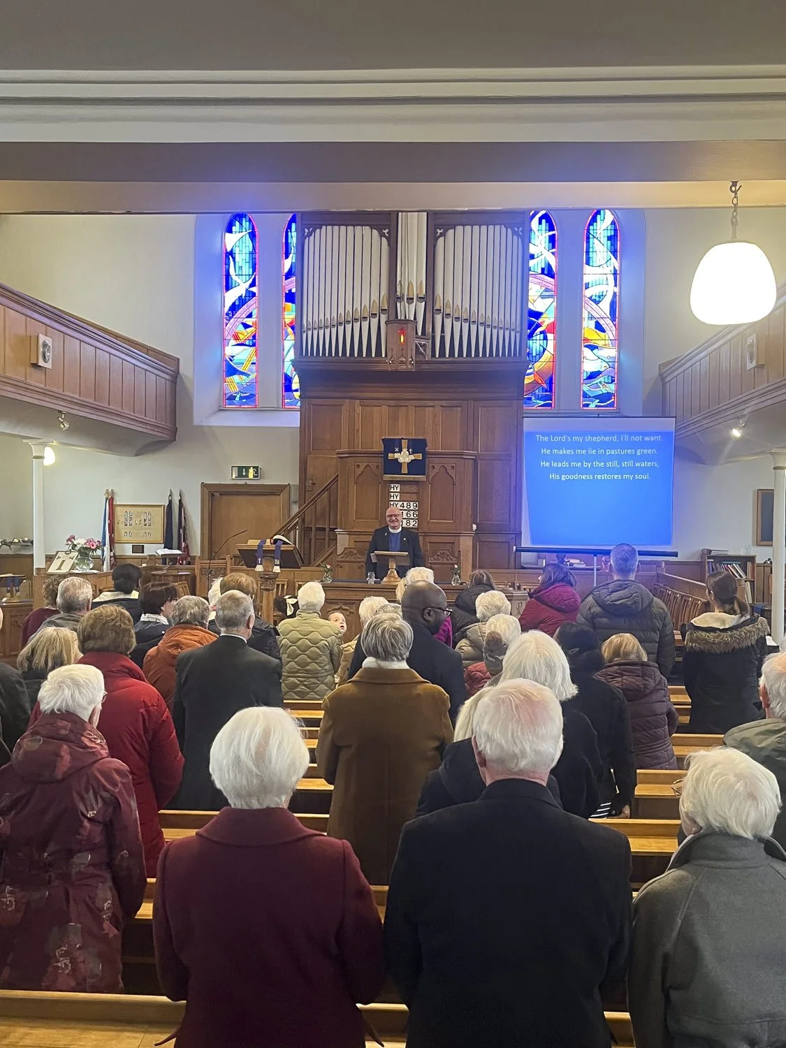 A church congregation gathered for a service, with a minister at the pulpit and stained glass windows behind him, in a church with wooden fixtures and a screen displaying lyrics.