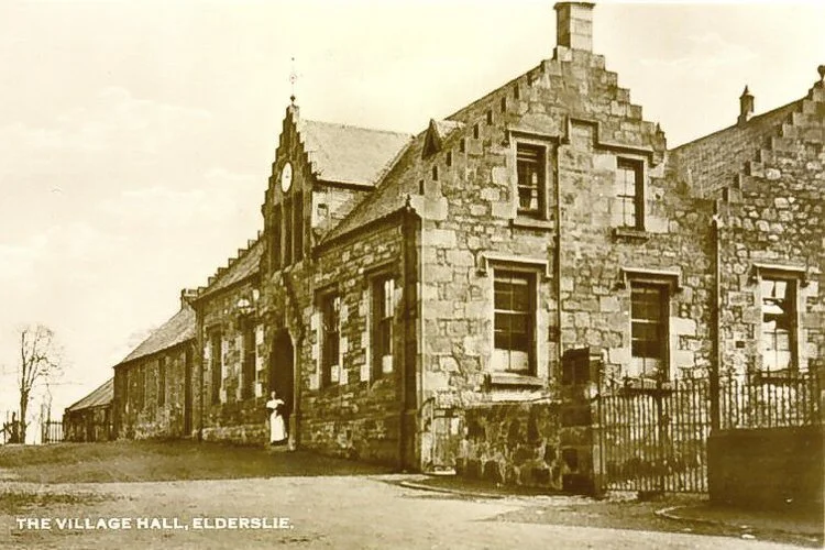 A sepia-toned vintage photograph of the village hall in Elderslie, a stone building with a stepped gable and a large clock on the front facade, and a woman in period clothing standing near the entrance.