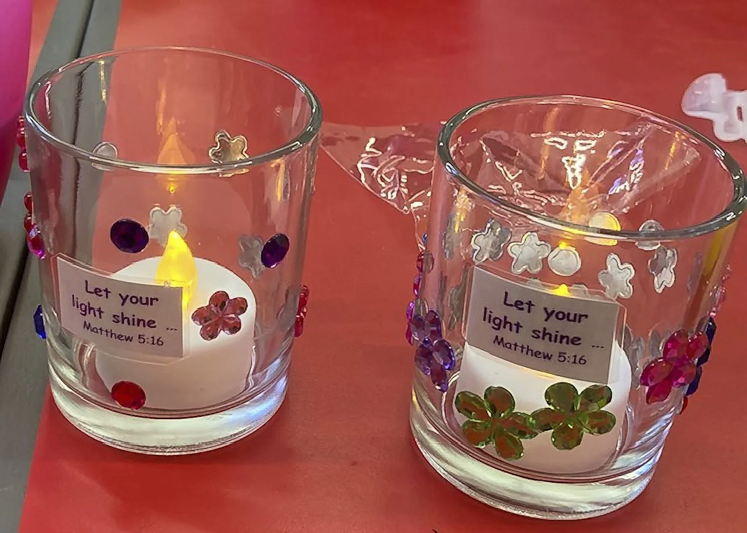 Two glass candle holders decorated with colorful beads and flowers, each with a lit candle inside, placed on a red table. The candle holders have a label with the Bible verse: 'Let your light shine.' Matthew 5:16