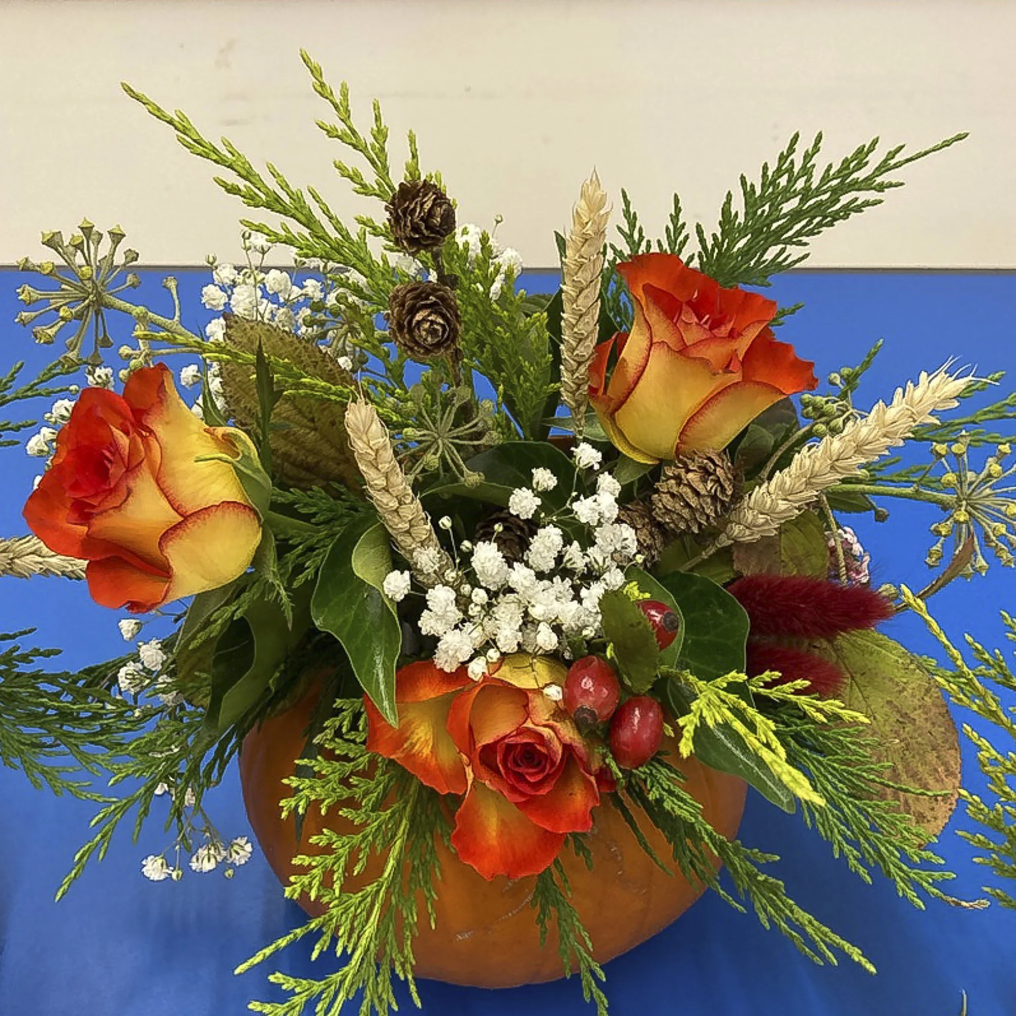 An autumn-themed floral arrangement with orange and yellow roses, white baby's breath, pine cones, red berries, green pine, and other foliage in a pumpkin-shaped container on a blue table.