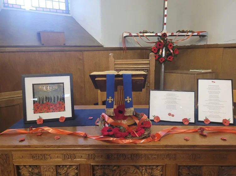 Communion table with display of framed photos and documents, and a wooden cross adorned with red poppies and ribbons, commemorating fallen soldiers.