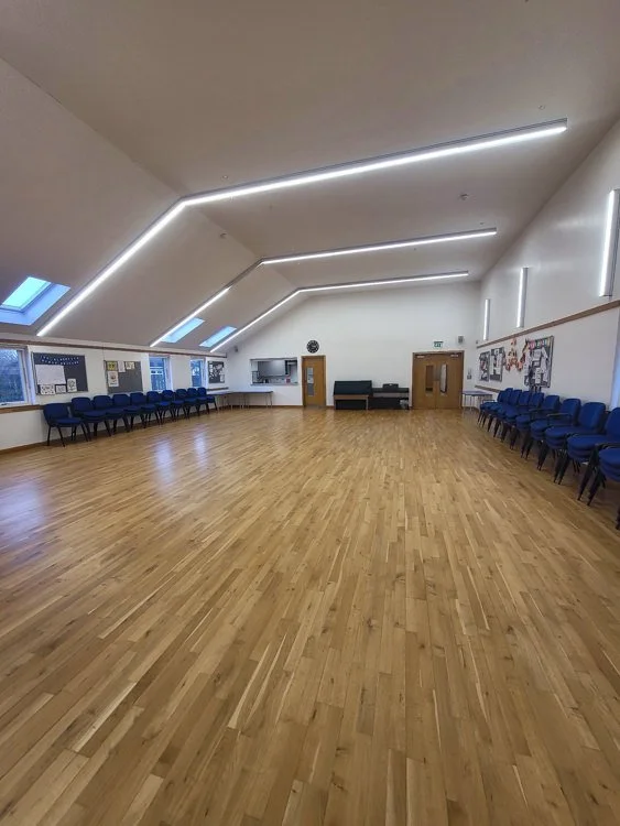 Large church hall with wooden floor, blue chairs along walls, bulletin boards, and skylights in the ceiling.