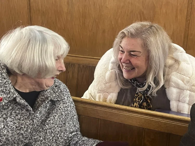 Two women talking and smiling in a wooden a wooden church pew.