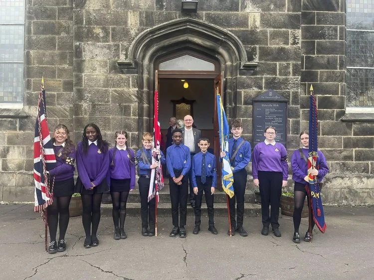 A group of students and an adult standing with flags outside a historic stone building with an arched doorway.